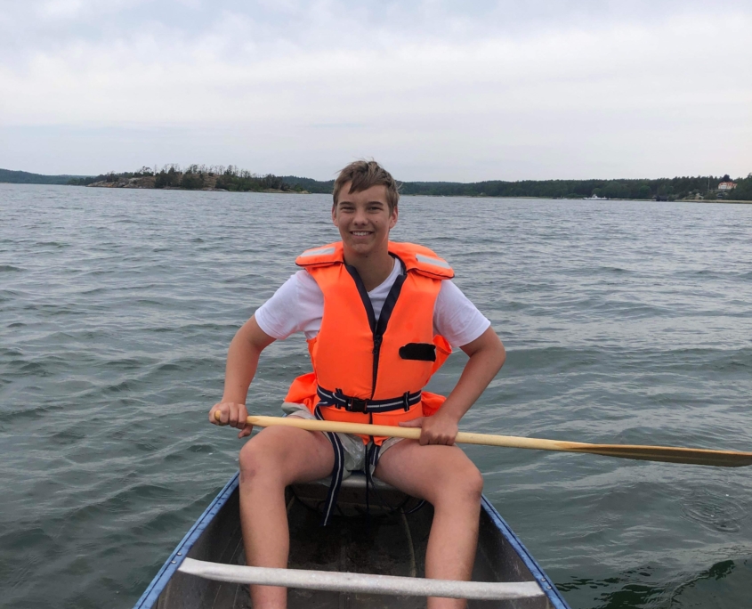 Pojke med kort mellanmörkt hår sitter i en roddbåt och håller i en paddel på vatten.